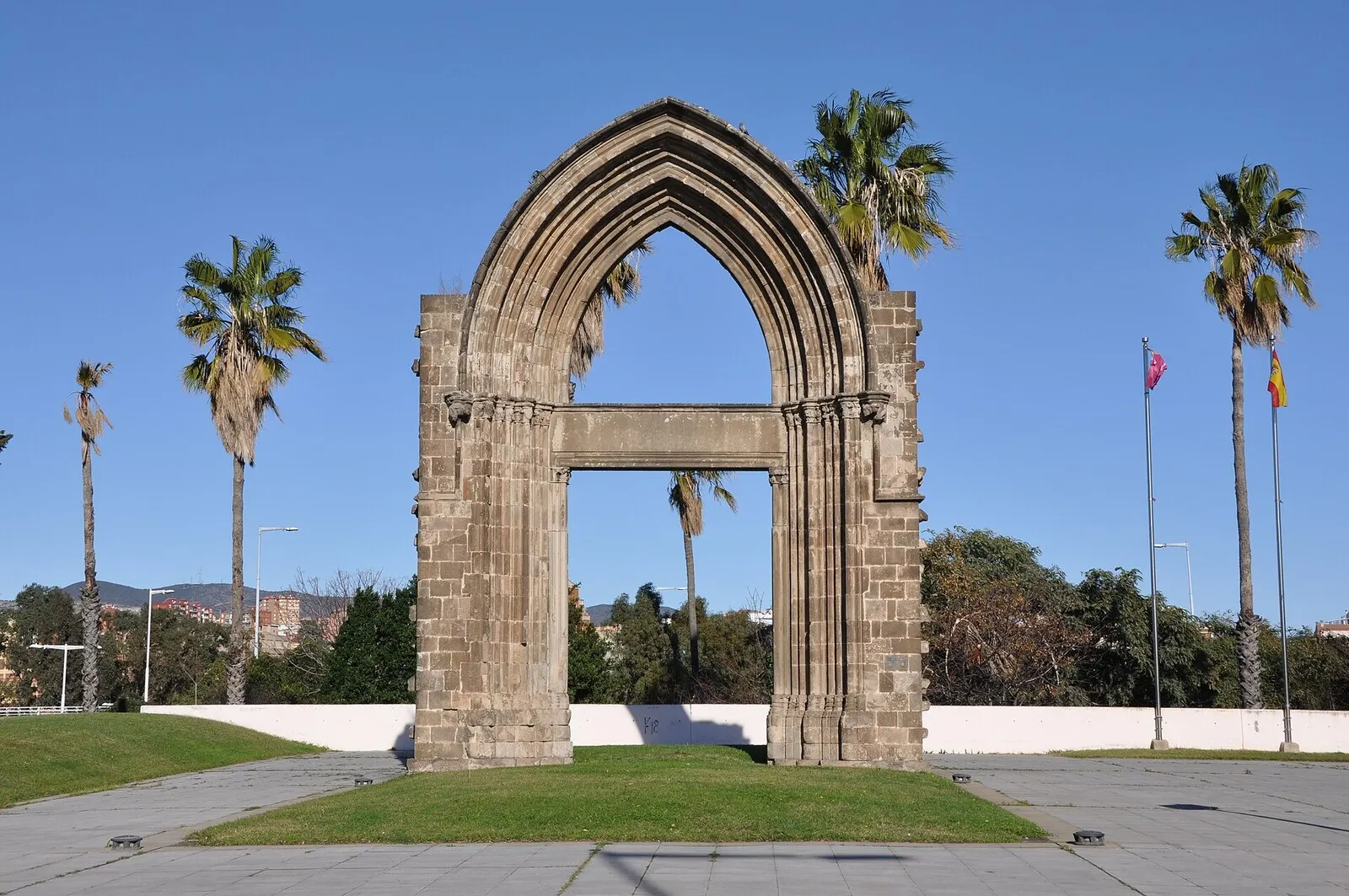 Un gran arco gótico de piedra aislado sobre el césped, entre palmeras y bajo un cielo azul, en Sant Adrià de Besòs