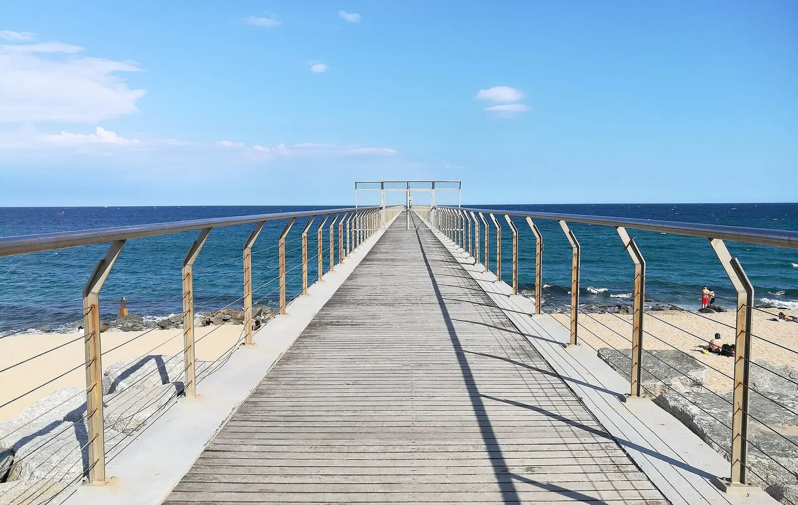 La pasarela de madera del Pont del Petroli adentrándose en el mar desde la playa de Badalona, con cielo azul