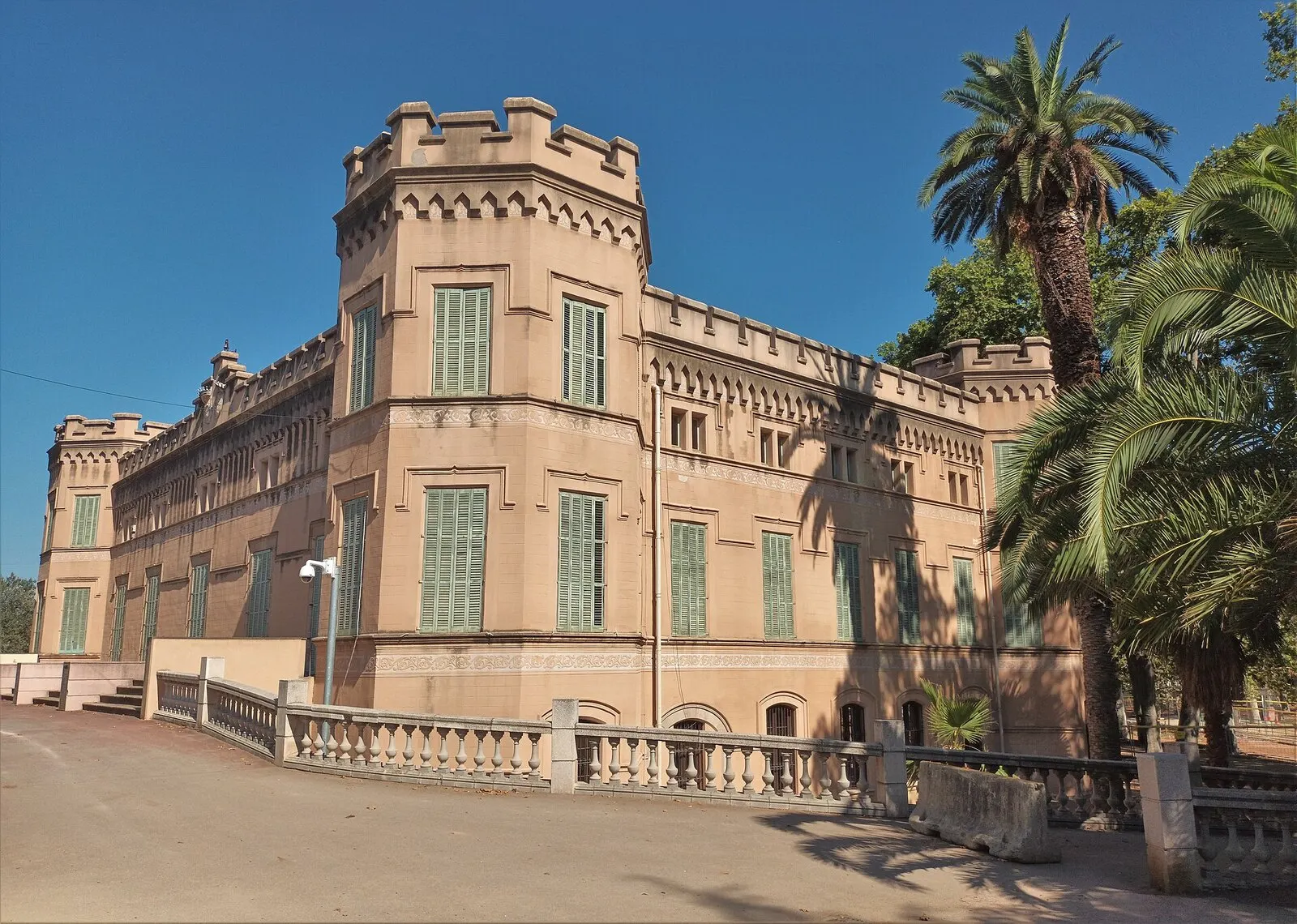 Palacete de Can Mercader en Cornellà, con torre almenada y persianas verdes, entre palmeras y una balaustrada