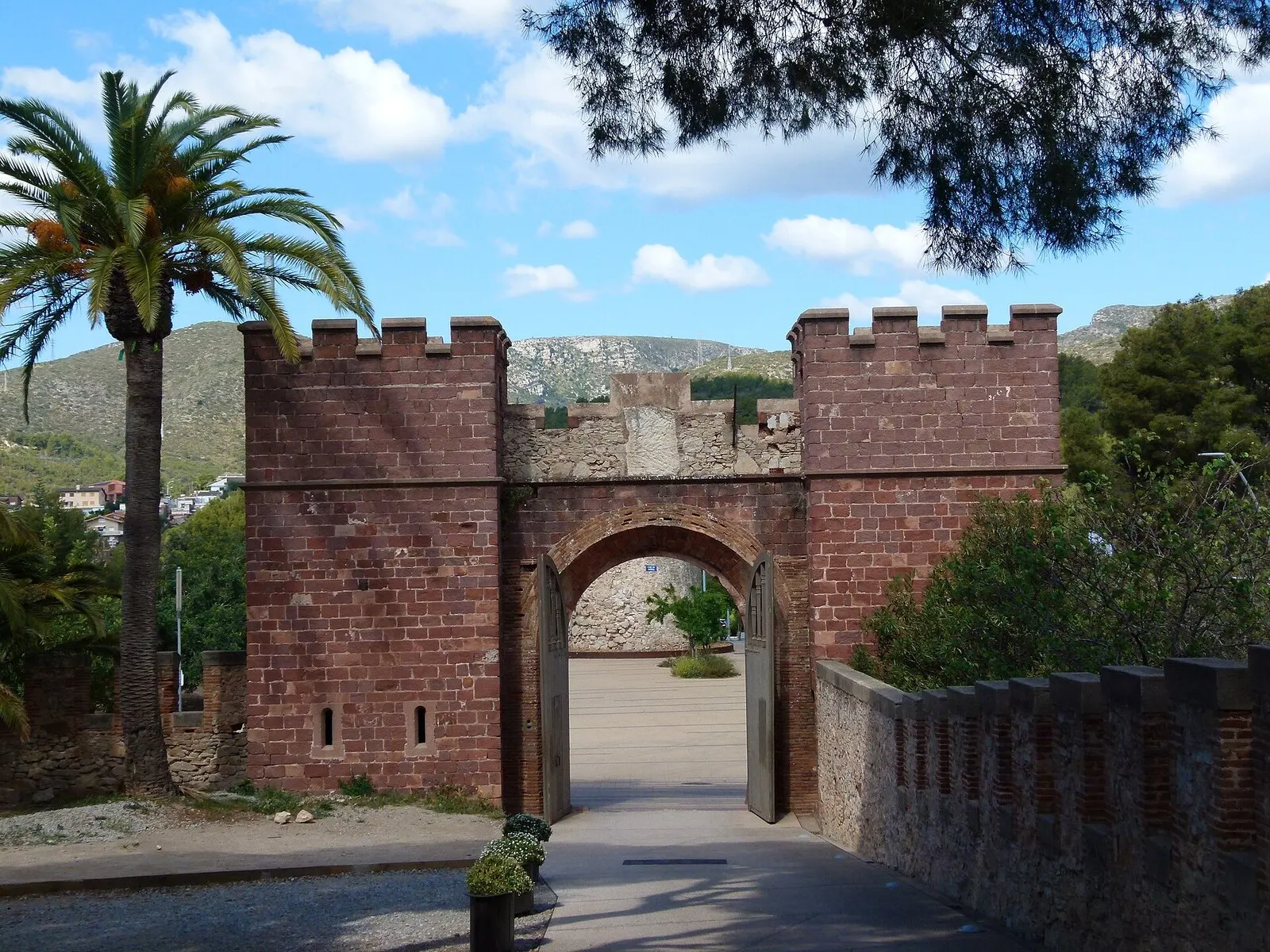 Puerta almenada de piedra rojiza del castillo de Castelldefels, entre una palmera y un pino, con montañas detrás