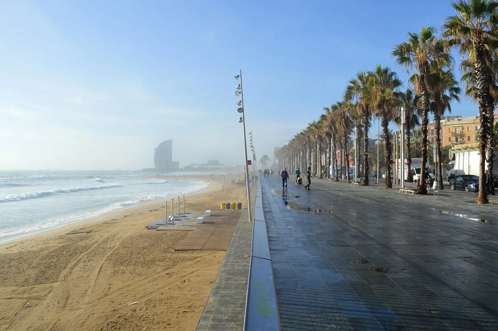 Paseo marítimo de la Barceloneta con palmeras junto a la playa, y el hotel W entre la bruma de la mañana al fondo