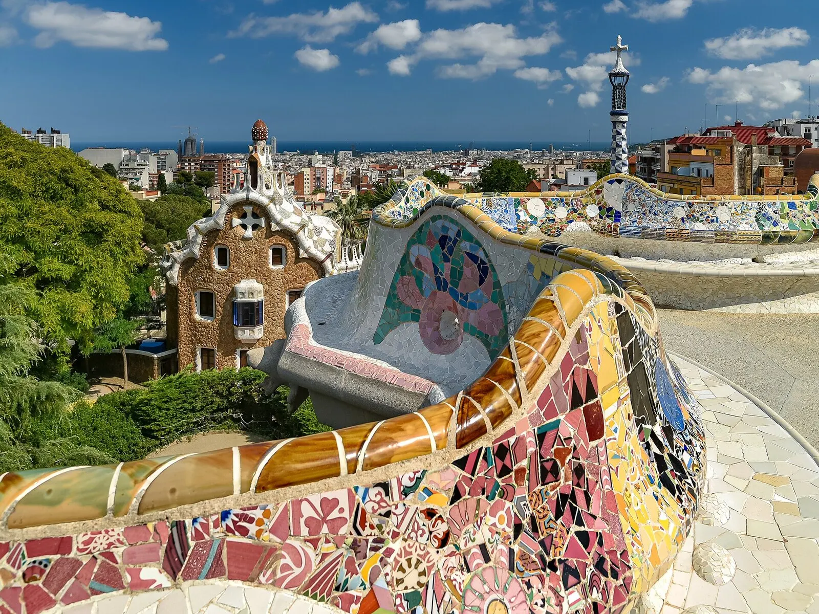 Banco ondulado de trencadís del Park Güell con un pabellón de la entrada detrás y Barcelona hasta el mar al fondo