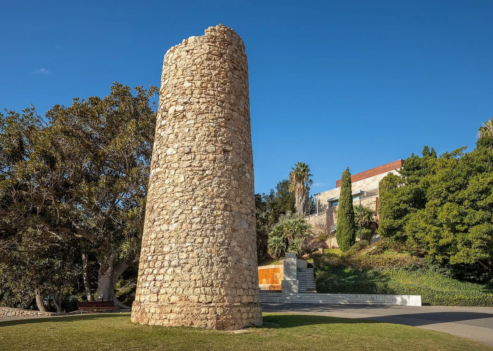 La Torre de la Linterna, una atalaya cilíndrica de piedra, entre los árboles del Parque Torres con cielo azul