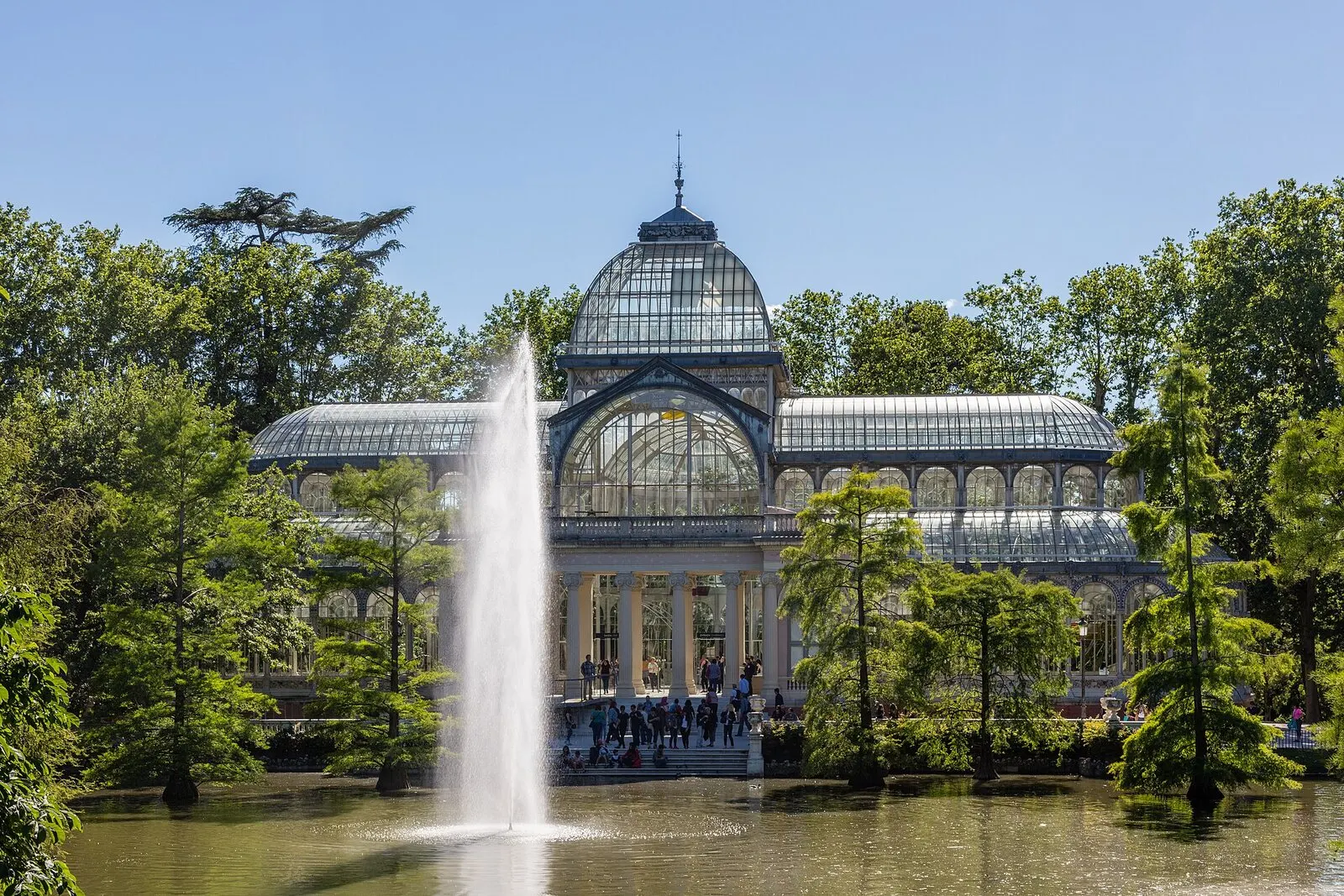 El Palacio de Cristal del Retiro tras su estanque, con un surtidor y árboles alrededor, en un día soleado