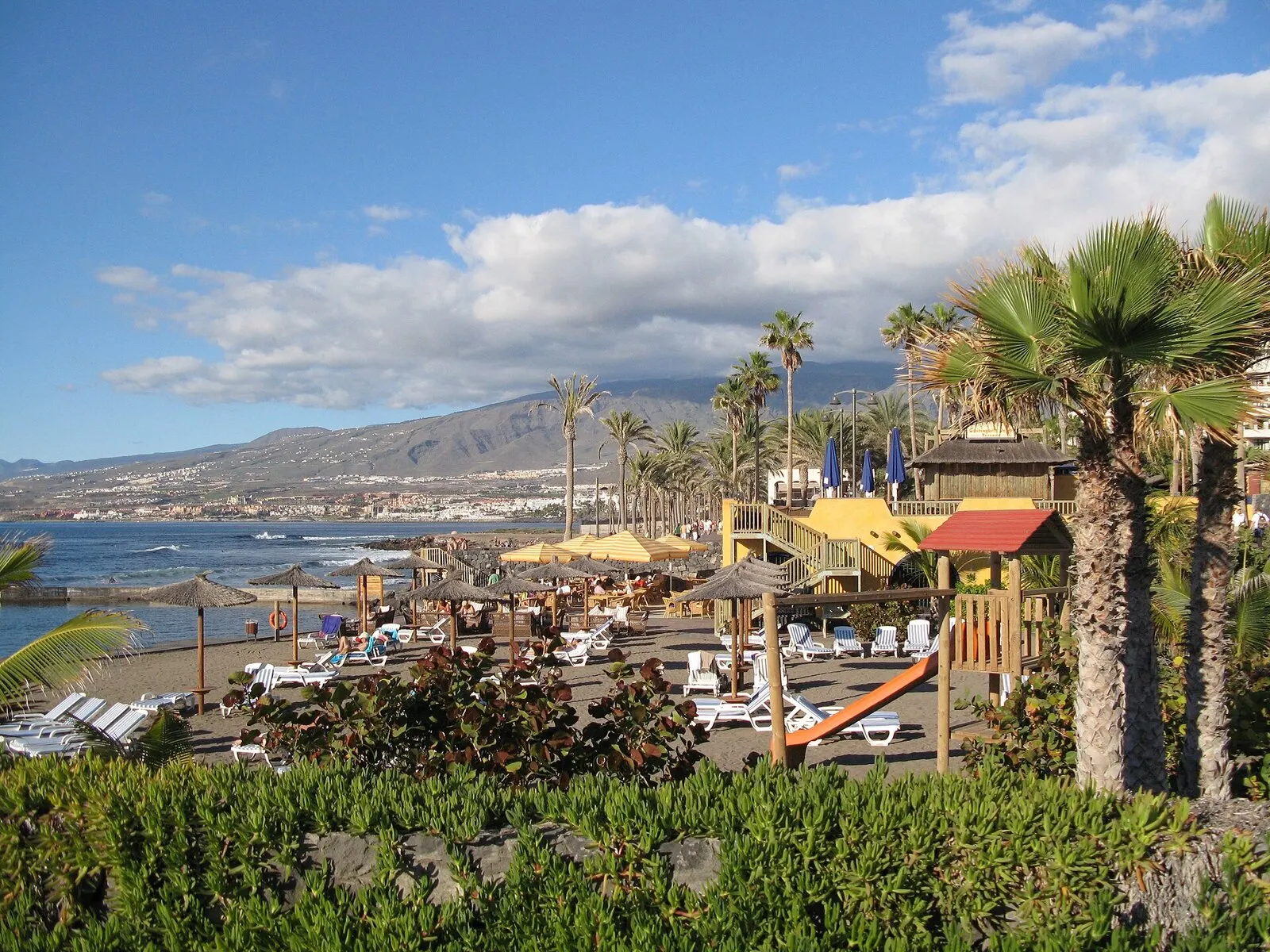 Playa de arena oscura de Las Américas con sombrillas de paja, palmeras y las laderas del sur de Tenerife detrás