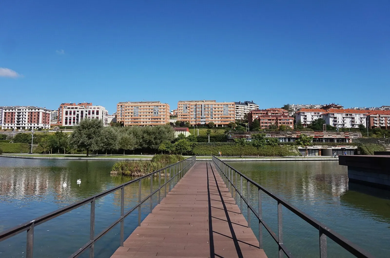Pasarela de madera sobre el lago del parque de Las Llamas, con edificios de viviendas al fondo