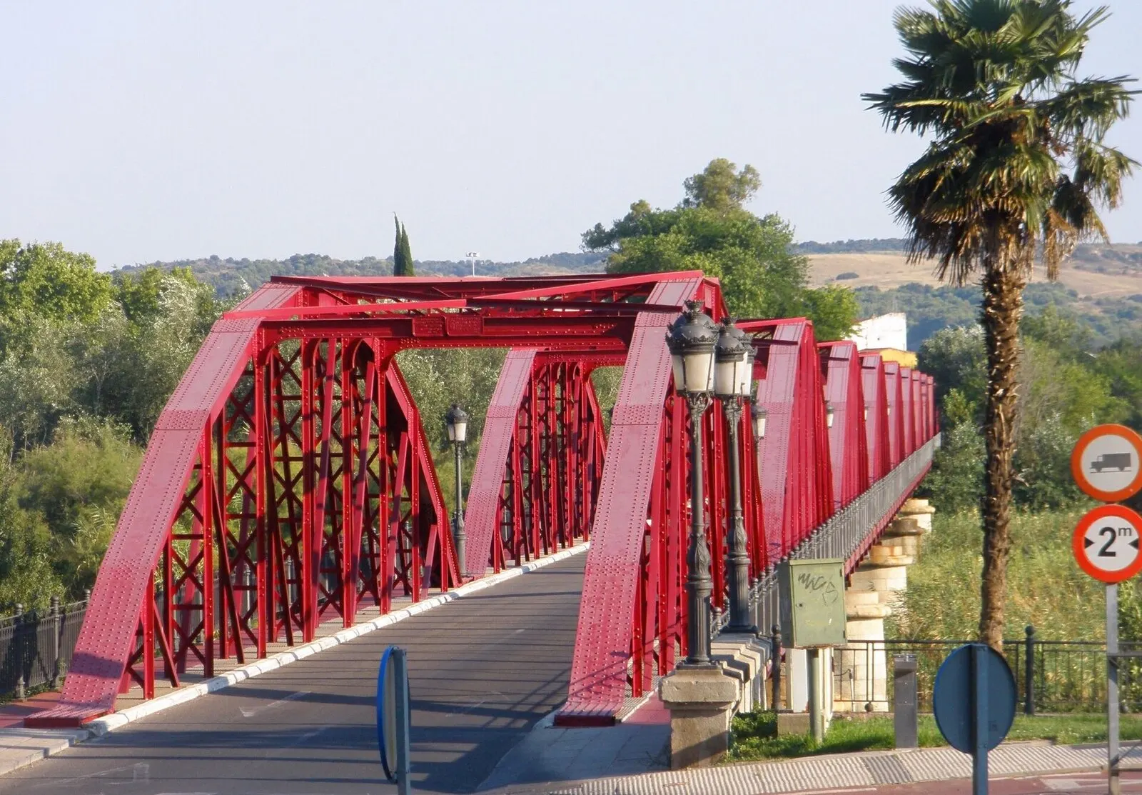 Los arcos rojos del puente de Hierro sobre el Tajo, con farolas y vegetación de ribera al fondo