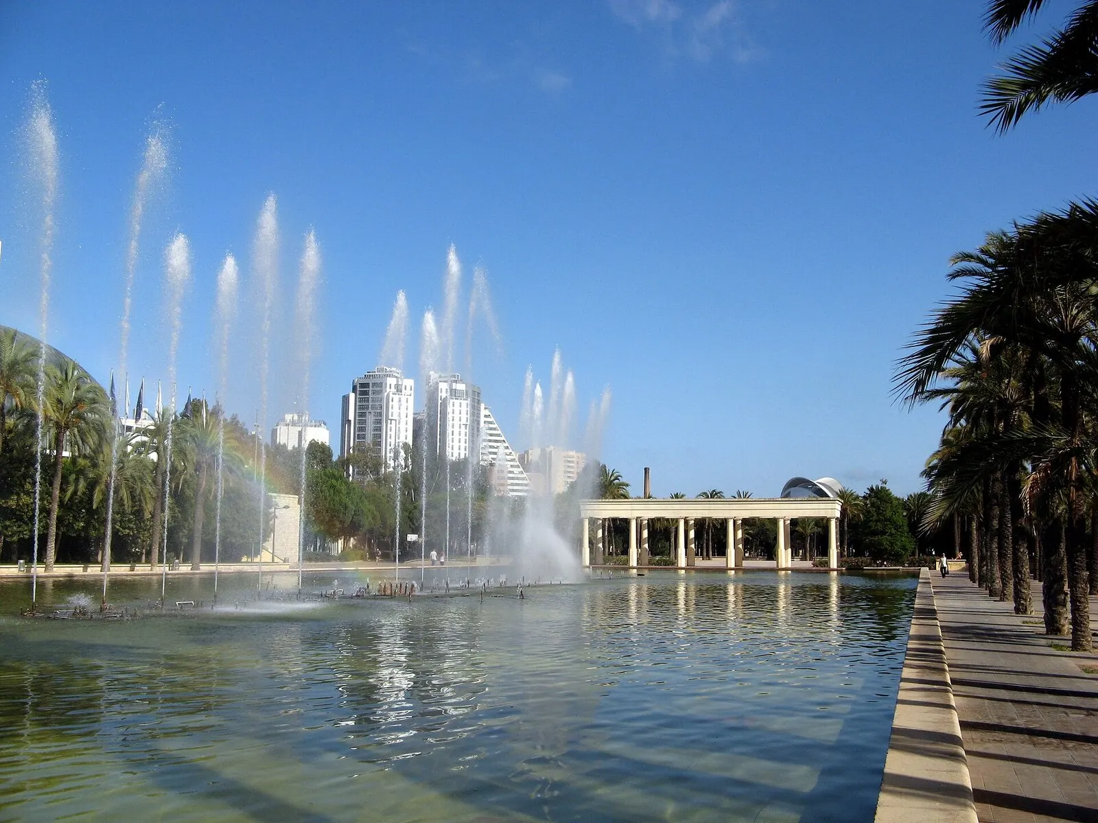 Un estanque con surtidores en el Jardín del Turia, con una pérgola y edificios altos al fondo, en un día soleado